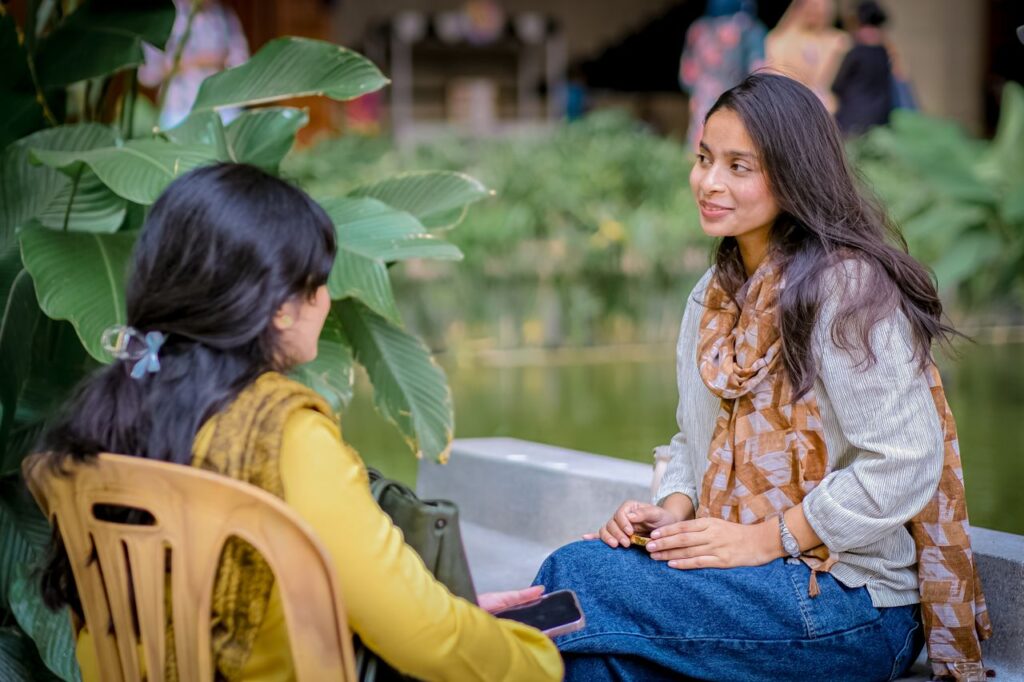 Two women engaged in conversation by a garden pond, surrounded by lush greenery.